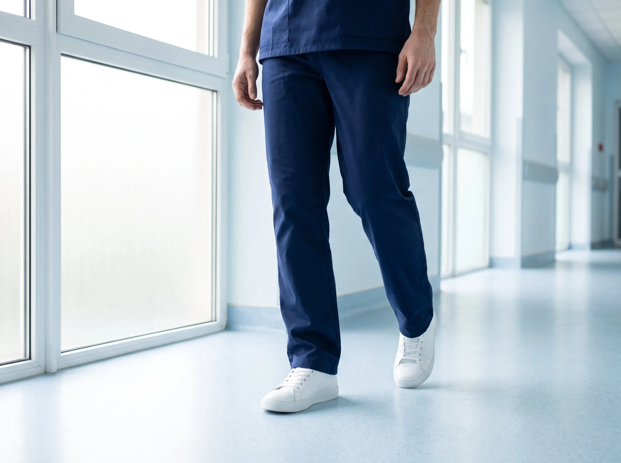 Person in navy scrubs and white trainers walking along a bright hospital corridor